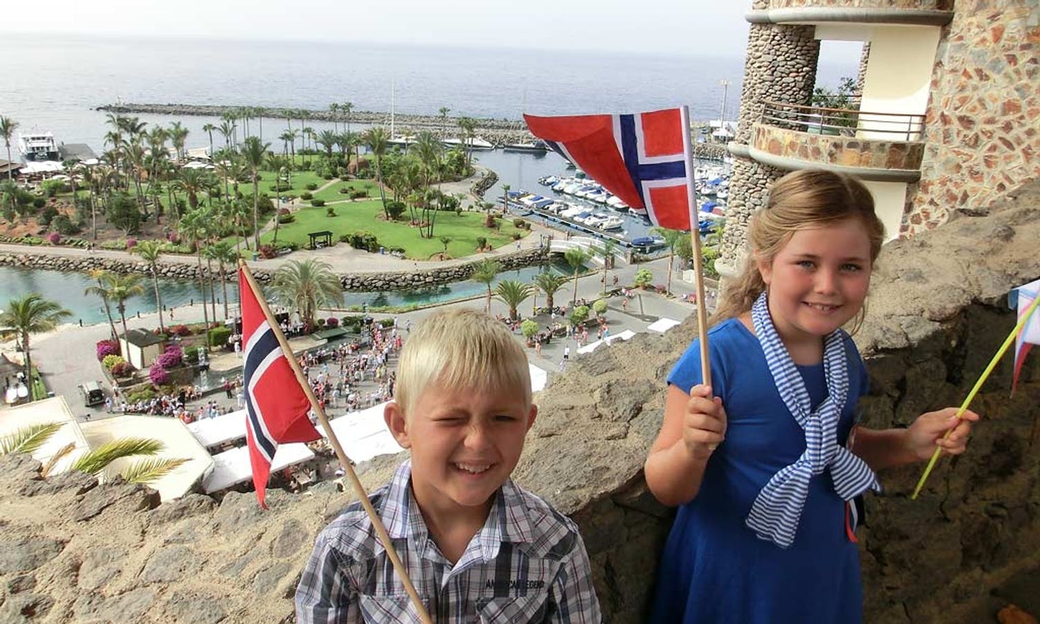 Petter(6) og Sofia (7) veivar med norske-flagga og synst 17.mai på Gran Canari er kjempekult. Kan tru alle turistane som sperra opp augene når "toget" passerte stranda. Her med hjerteøya og tale på Anfi-plaza i bakgrunnen. (privat foto)